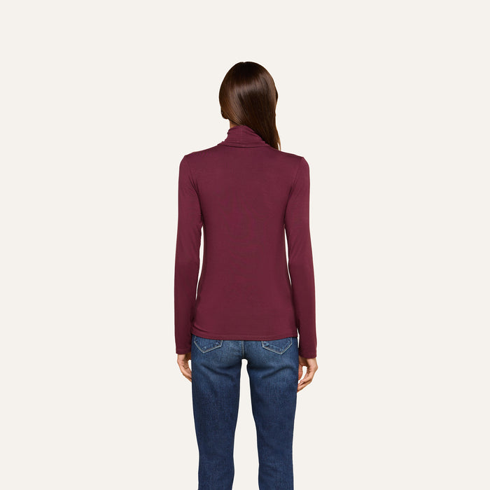 A woman stands facing away, wearing the slim fit Lani Turtleneck in maroon paired with blue jeans against a plain, light background—a perfect look for Fall 2026.