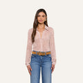 Woman with long brown hair wearing the Laurent Blouse—a light pink button-up top with a subtle floral lace print—paired with blue jeans and a tan belt, standing against a plain light background.
