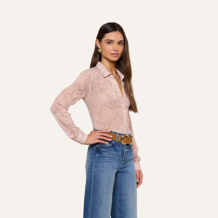 A woman with long brown hair wears the Laurent Blouse—a relaxed fit, long sleeve, light pink patterned top—paired with blue jeans, a brown belt, and gold earrings. She stands with one hand on her hip against a plain light background.