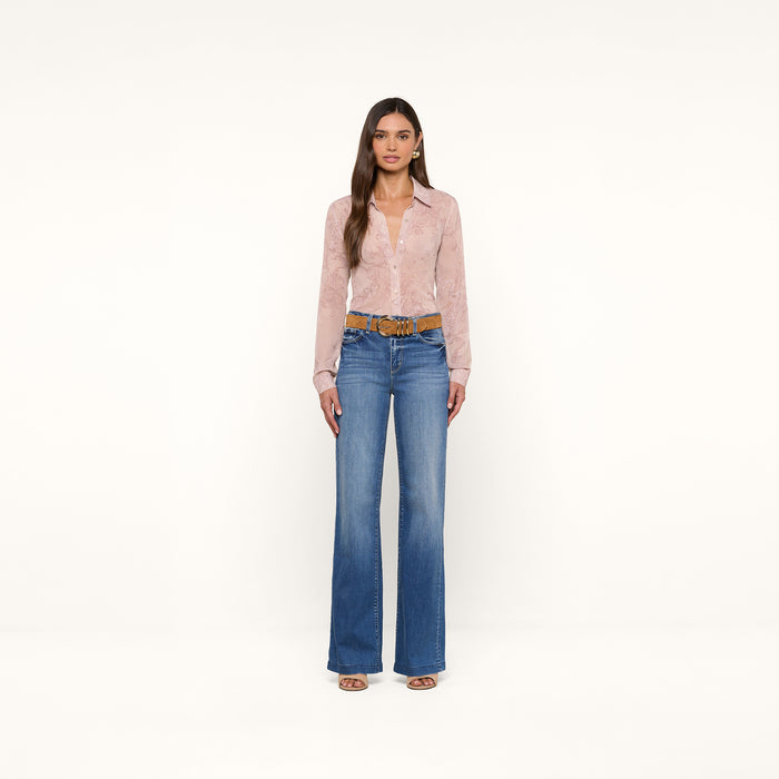 A woman stands against a plain white background, wearing the Laurent Blouse—a light pink, sheer poly chiffon button-up shirt with long sleeves and a relaxed fit—paired with blue high-waisted wide-leg jeans, a brown belt, and open-toe shoes.