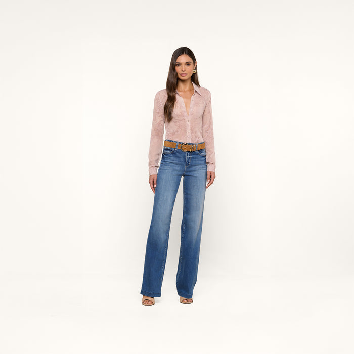 A woman stands against a plain white background, wearing the Laurent Blouse—a light pink, relaxed-fit long sleeve top in sheer poly chiffon—paired with blue high-waisted wide-leg jeans, a brown belt, and open-toed shoes.
