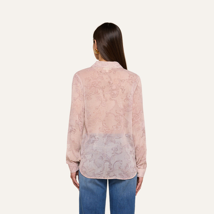 A woman with straight brown hair is shown from the back against a plain background, wearing the Laurent Blouse—a light pink, sheer poly chiffon blouse with a relaxed fit, long sleeves, and a subtle pattern—paired with blue jeans.