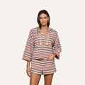 Model wears the Lee Cover-Up Poncho—a vibrant, striped, long-sleeve hooded top with a lace-up neckline and matching shorts—perfect for après-swim or poolside lounging. She stands facing forward with one hand in her shorts pocket against a plain background.