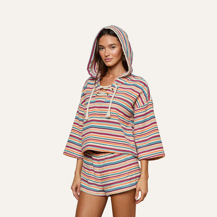 A woman wearing the Lee Cover-Up Poncho—a hooded, long-sleeve terrycloth poncho with matching shorts featuring colorful horizontal stripes—stands against a plain light background, making it an ideal choice for poolside lounging or après-swim relaxation.