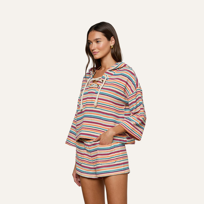 Wearing the Lee Cover-Up Poncho—a colorful, striped hoodie with a lace-up front and matching shorts—this woman stands with one hand in her pocket against a plain light background, ready for relaxed poolside lounging.
