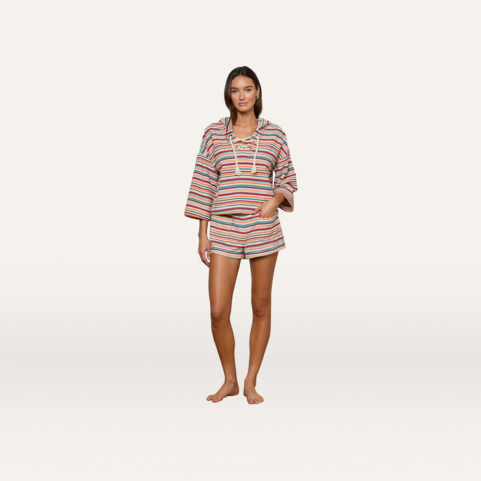 A woman stands barefoot against a plain white background, wearing the Lee Cover-Up Poncho—a colorful striped long-sleeved top with a lace-up neckline and matching shorts—perfect for après-swim or poolside lounging.