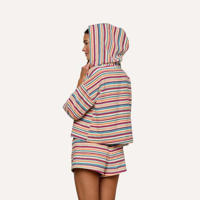 Woman in the Lee Cover-Up Poncho—a colorful, striped terrycloth hooded top with matching shorts—stands sideways against a plain background, her hood up and hand touching her chin. Perfect for après-swim or poolside lounging.