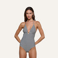 A woman stands with hands on hips against a plain background, exuding French glamour in the Leila Halter One-Piece Swimsuit—a black-and-white striped design featuring a deep V-neck and halter straps.