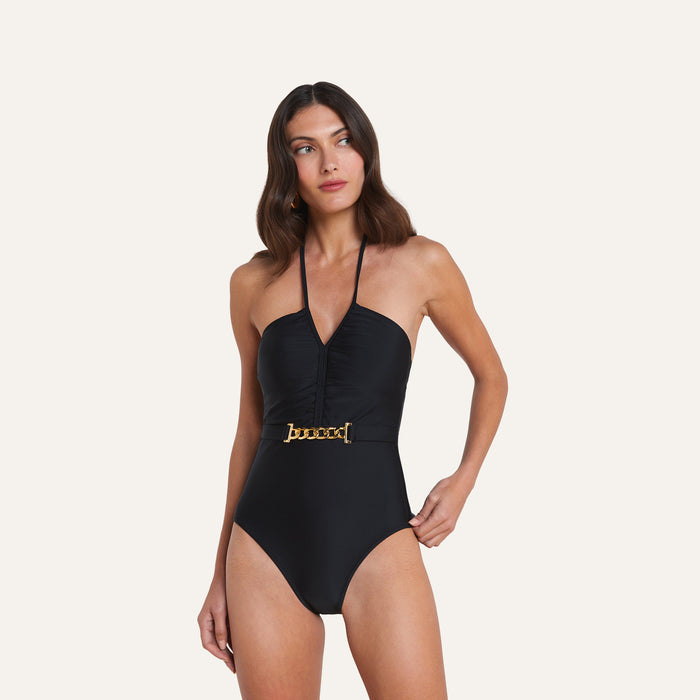 A woman wears the Leila Halter One-Piece Swimsuit, featuring an adjustable halter neckline, deep V-cut, and a gold chain accent at the waist, as she stands against a plain light background.