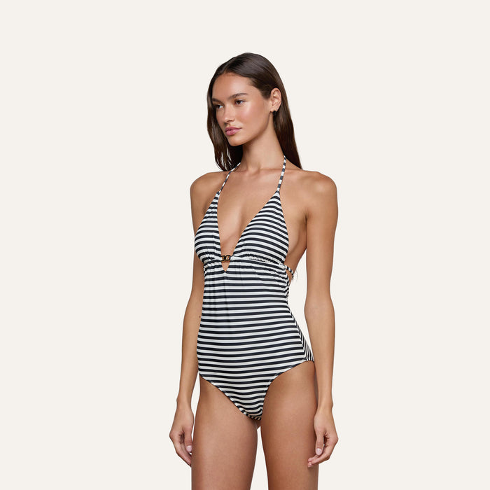 A woman models the Leila Halter One-Piece Swimsuit in a black-and-white stripe pattern, standing against a plain light background and gazing slightly to the side, capturing subtle French glamour.