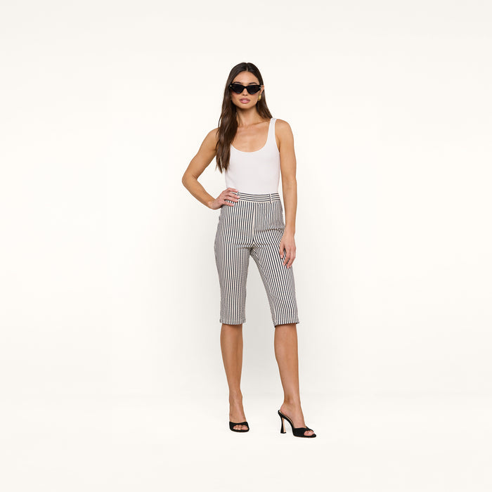 A woman stands confidently against a plain white background, wearing a white tank top, the Leni Pedal Pusher in black-and-white striped cotton seersucker, black high heels, and sunglasses.