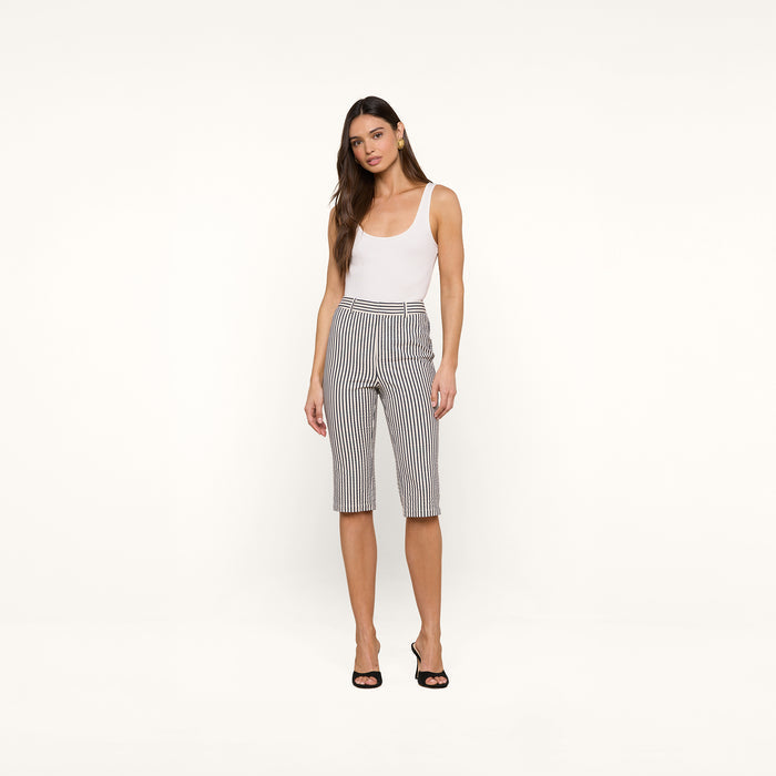 A woman stands against a white background, wearing a white tank top, the Leni Pedal Pusher in black and white vertical striped cotton seersucker, and black open-toe heels.