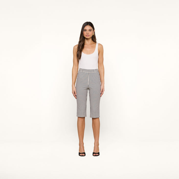 A woman stands against a plain white background, wearing a white tank top, the Leni Pedal Pusher high-waisted knee-length black-and-white cotton seersucker shorts, and black open-toe heels.