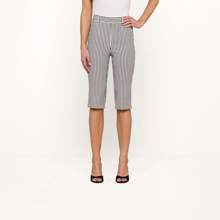 A person stands against a plain background, showcasing the lower half of their body dressed in the Leni Pedal Pusher—black and white vertically striped cotton seersucker knee-length shorts—paired with a sleeveless white top and black open-toe heels.