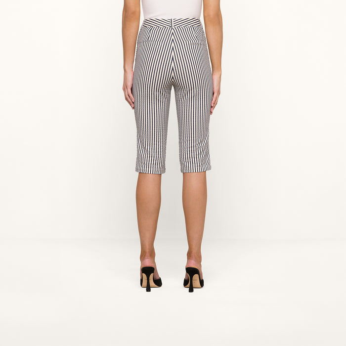 A person stands facing away against a plain white background, wearing the Leni Pedal Pusher—knee-length black and white vertical striped cotton seersucker shorts—paired with a white top and black open-heeled shoes.