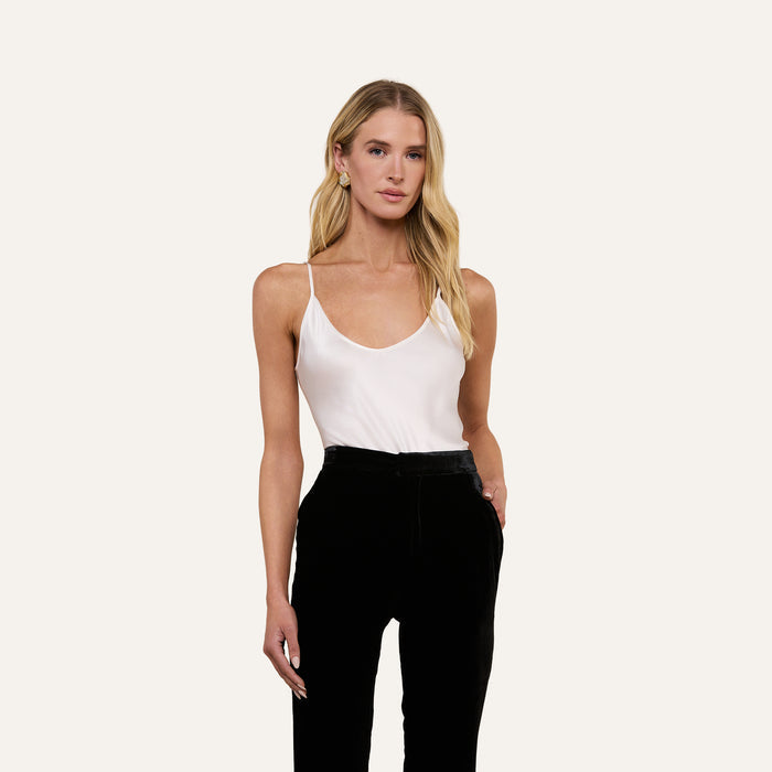 Wearing the Lexi Silk Camisole in white with adjustable straps and black pants, a woman stands with one hand in her pocket against a plain light background.