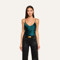 A woman stands against a plain light background, facing forward with relaxed arms at her sides, wearing the Lexi Silk Camisole in dark green—featuring a bias-cut silhouette and adjustable straps—paired with black leather pants accented by a wide belt with a gold buckle.