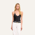 A woman with long blonde hair stands against a plain light background, wearing the Lexi Silk Camisole with adjustable spaghetti straps, paired with white pants, a black belt, and earrings.