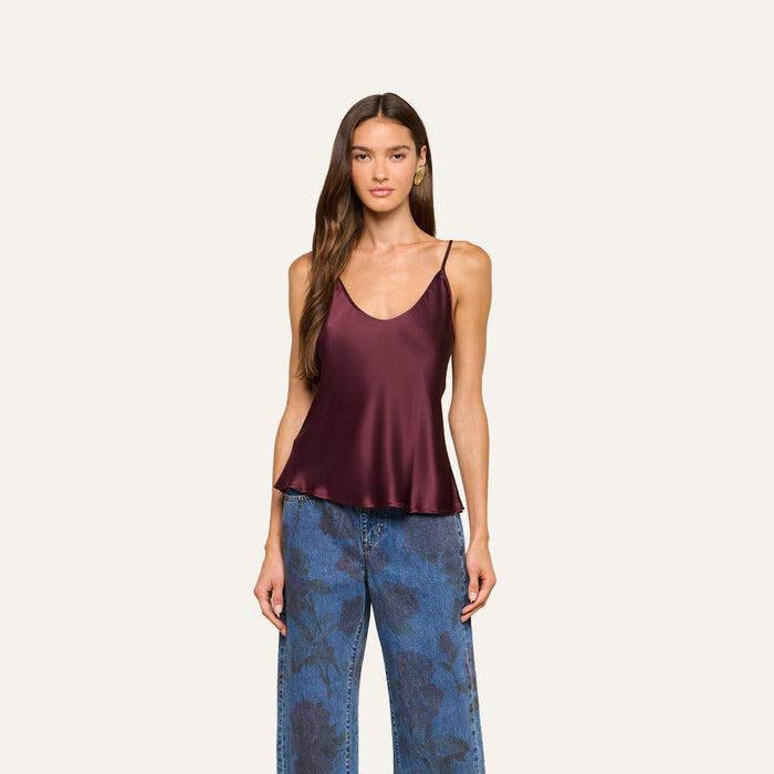 A woman wearing the Lexi Silk Camisole, a sleeveless deep burgundy bias-cut silk top with adjustable straps, paired with high-waisted blue jeans featuring a floral pattern, stands against a plain light background.