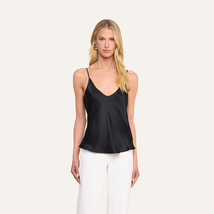 A woman stands against a plain light background, facing forward with her arms at her sides, wearing the Lexi Silk Camisole in black with adjustable spaghetti straps and white pants.