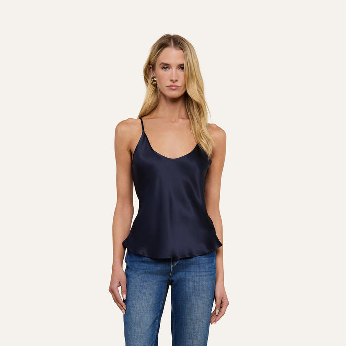A woman with long blonde hair, wearing the Lexi Silk Camisole in midnight blue and blue jeans, stands against a plain light background, looking at the camera.