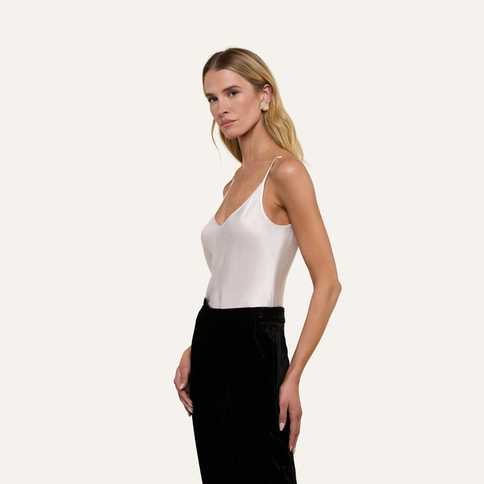 A woman with long blonde hair wears the Lexi Silk Camisole, a sleeveless white charmeuse top with adjustable straps, paired with black pants. She stands against a plain light background, looking slightly to the side.