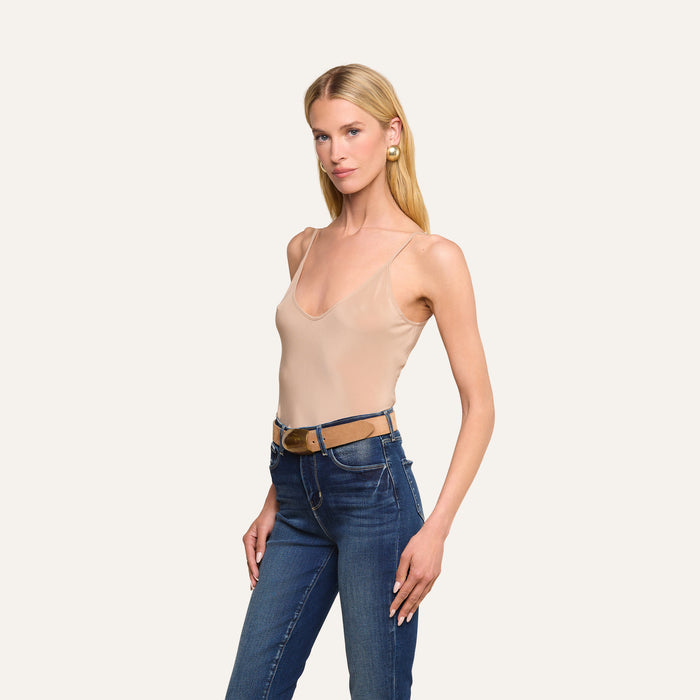 A woman with long blonde hair, wearing the Lexi Silk Camisole—a beige tank top with adjustable straps—paired with blue jeans, a brown belt, and large earrings, stands against an off-white background, posing with one hand on her hip.