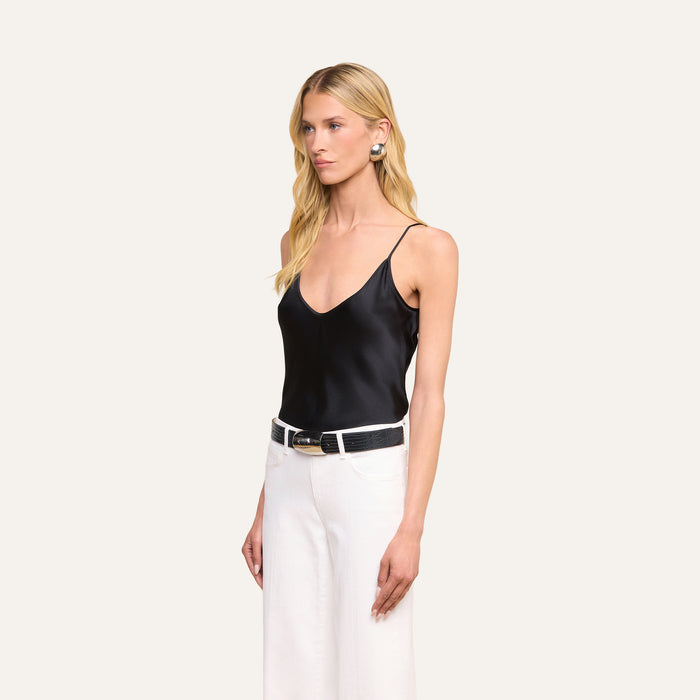 A woman with long blonde hair wears the Lexi Silk Camisole—a black silk top with adjustable spaghetti straps—paired with white high-waisted pants, a black belt, and statement earrings. She stands against a plain light background, looking slightly to her right with a neutral expression.