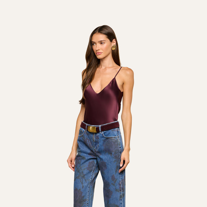 A woman with long brown hair wears the Lexi Silk Camisole in burgundy, featuring adjustable straps, paired with blue floral-patterned jeans and a brown belt, standing against a plain light background.