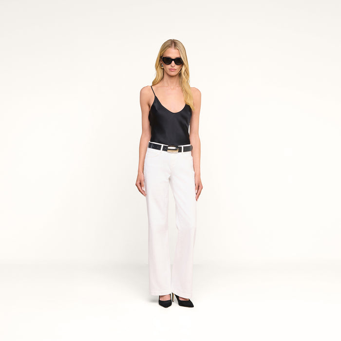 A woman wearing sunglasses, the Lexi Silk Camisole with adjustable spaghetti straps, white wide-leg pants, a black belt, and black heels stands against a plain white background.