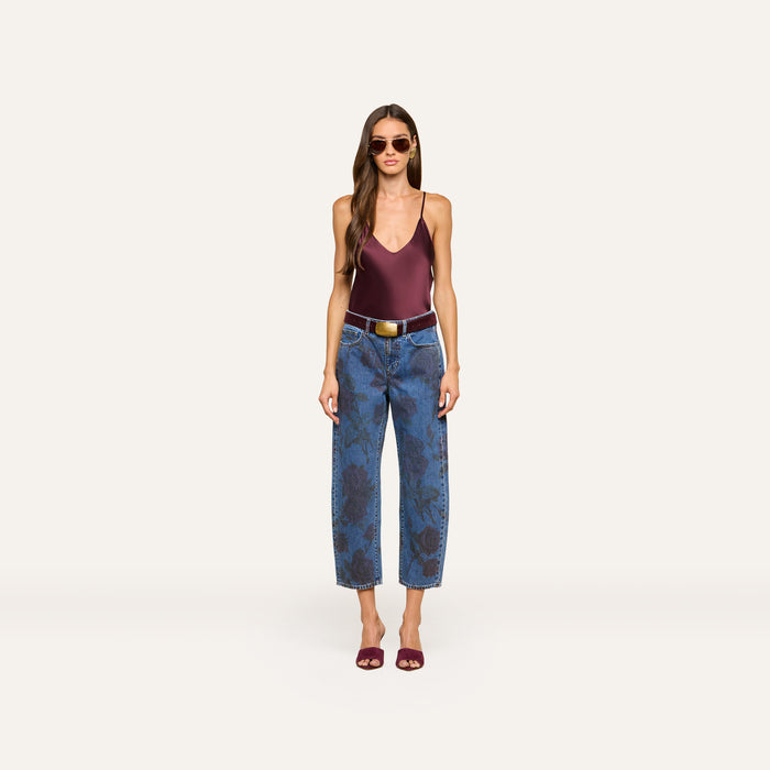 A woman wearing sunglasses, the Lexi Silk Camisole in maroon with adjustable straps, patterned blue jeans with a belt, and maroon heels stands against a plain light background.