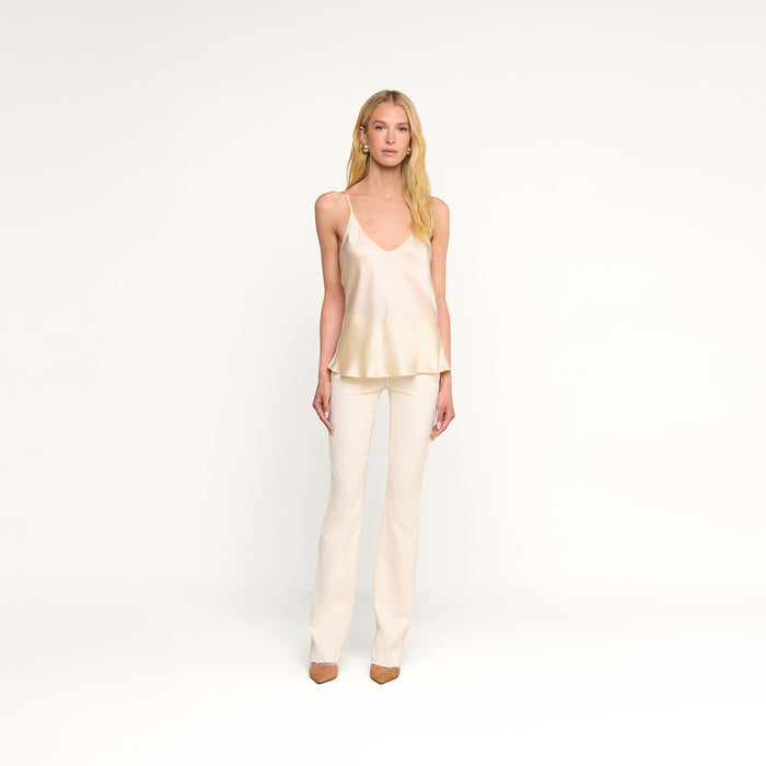 A woman stands against a plain white background, wearing the Lexi Silk Camisole in pearl white, paired with white pants and brown heels. She has long blonde hair and a neutral expression.