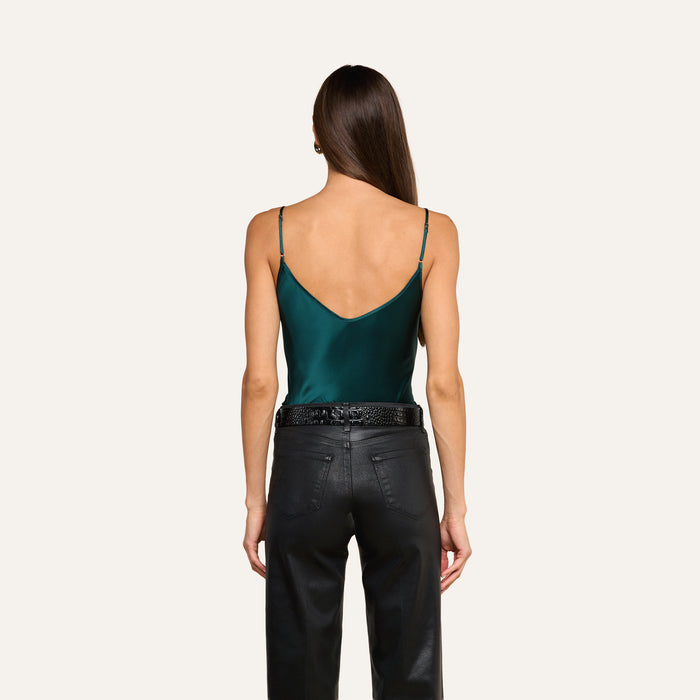 A woman stands with her back to the camera, wearing the Lexi Silk Camisole in dark green—with adjustable straps—and black high-waisted pants. Her straight brown hair cascades down her back, set against a plain light beige background.