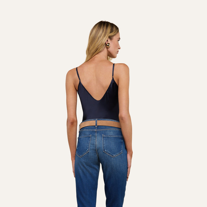 A woman with long blonde hair stands facing away, wearing the Lexi Silk Camisole in midnight blue with thin straps and blue jeans, set against a plain light background.