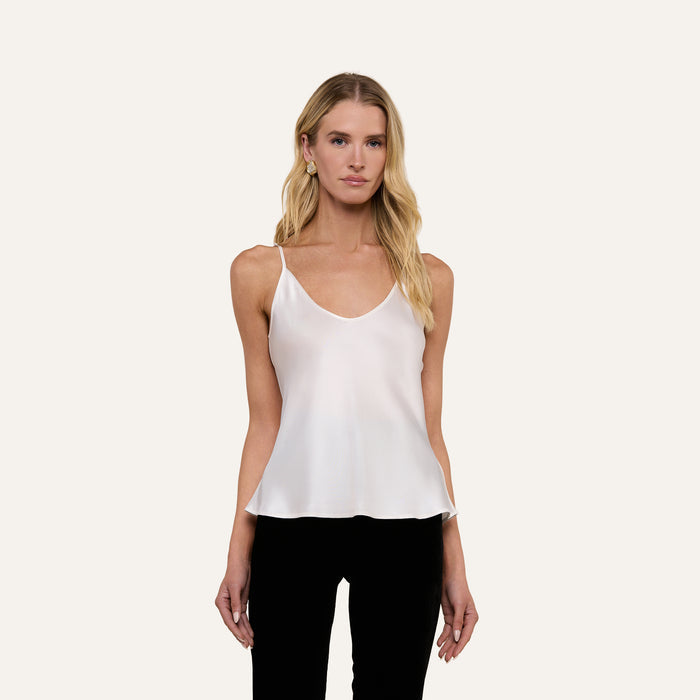 A woman with long blonde hair is wearing the Lexi Silk Camisole in white, featuring adjustable straps, paired with black pants as she stands facing forward against a plain light background.