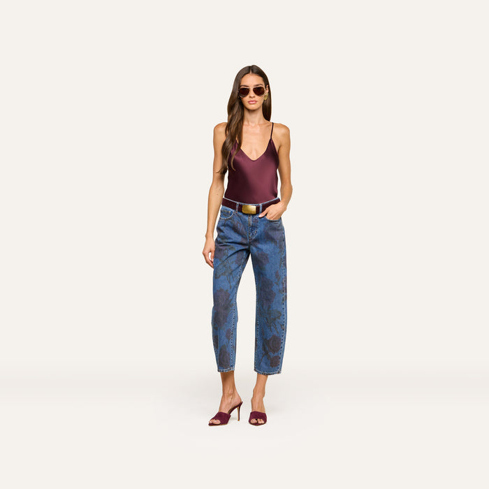 A woman wearing sunglasses, the Lexi Silk Camisole in maroon with adjustable straps, patterned blue jeans, and maroon heels stands confidently against a plain white background.