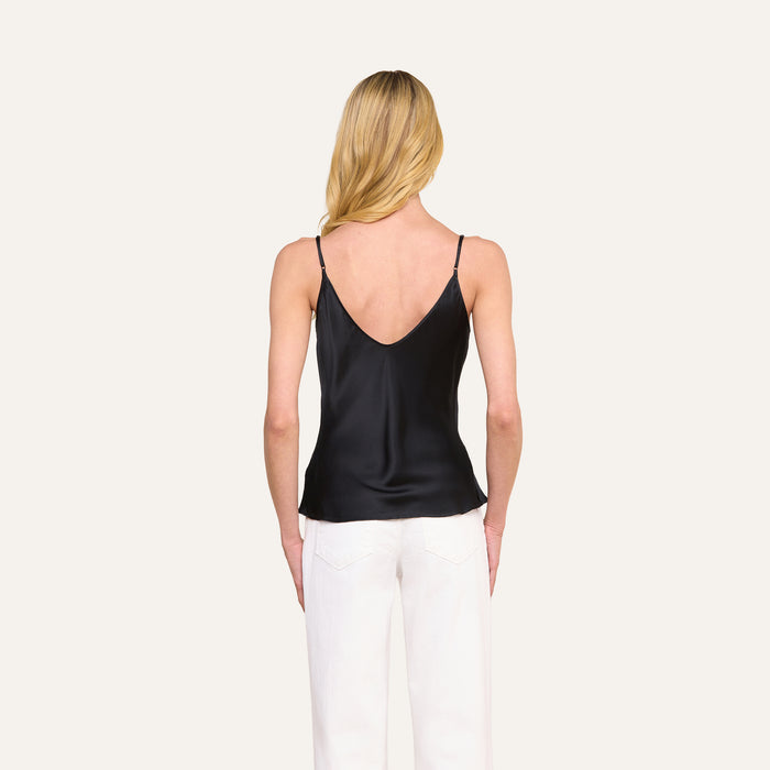 A woman with long blonde hair is seen from behind, standing against a plain light background, wearing the Lexi Silk Camisole in black with adjustable spaghetti straps and white pants.