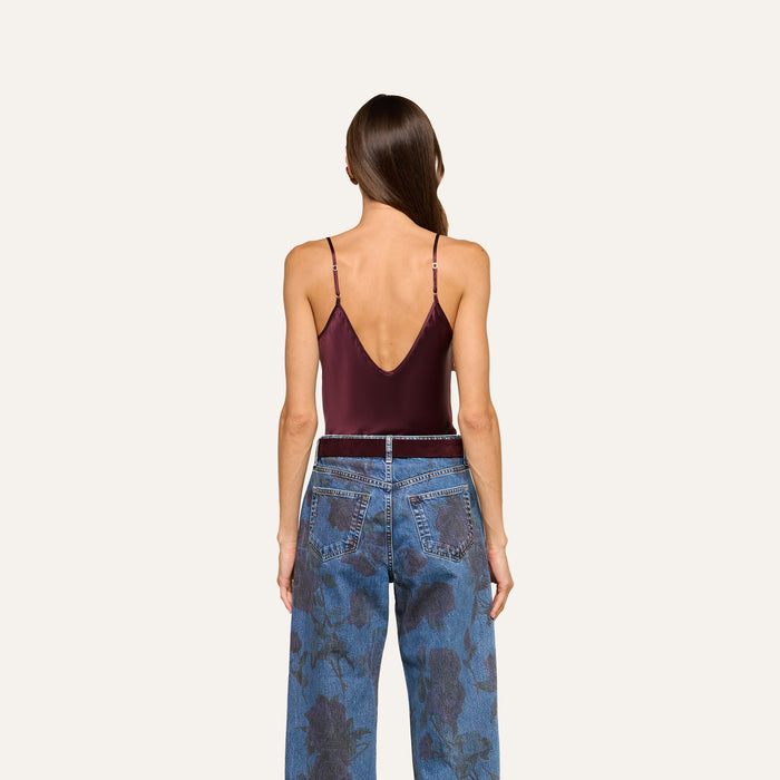 A woman stands with her back to the camera, wearing the Lexi Silk Camisole in burgundy with adjustable straps and high-waisted blue jeans featuring a floral pattern, set against a light, plain background.