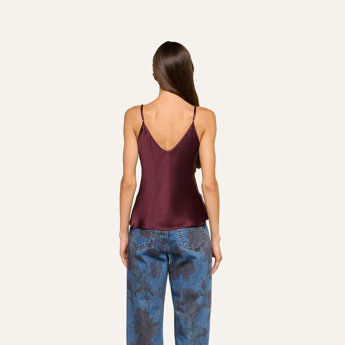 A woman with long brown hair is seen from behind, wearing the Lexi Silk Camisole—a sleeveless maroon top featuring a V-shaped back and adjustable straps—paired with blue floral-patterned jeans. The background is plain and light-colored.