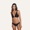 A woman with long brown hair models the Lexie Bikini Top, a black bikini featuring gold coin hardware on the top and adjustable straps, standing against a plain, light-colored background.