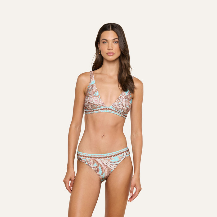 A woman stands wearing the Lexie Bikini Top, featuring a paisley print in light blue, tan, and cream hues. The adjustable straps provide added comfort and style. The background is plain and light-colored.