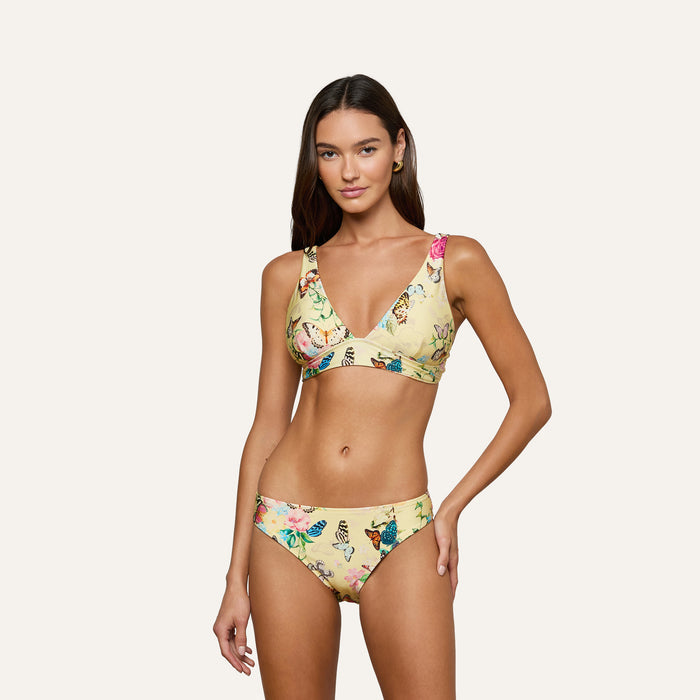 A woman poses in the Lexie Bikini Top, a yellow floral v-neck bikini featuring adjustable straps and matching bottoms, showcasing a colorful pattern against a plain light background.