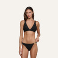 A woman models the Lexie Bikini Top in black jacquard terry, featuring a subtle floral pattern, as she poses against a plain light background with one hand resting on her bikini bottom.