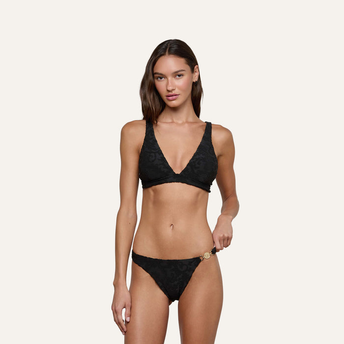 A woman models the Lexie Bikini Top in black jacquard terry, featuring a subtle floral pattern, as she poses against a plain light background with one hand resting on her bikini bottom.