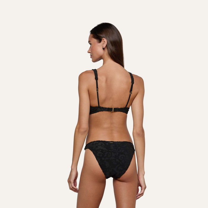A woman with long brown hair stands facing away, wearing the Lexie Bikini Top in black jacquard terry with adjustable straps and a gold clasp on the back, set against a plain light background.