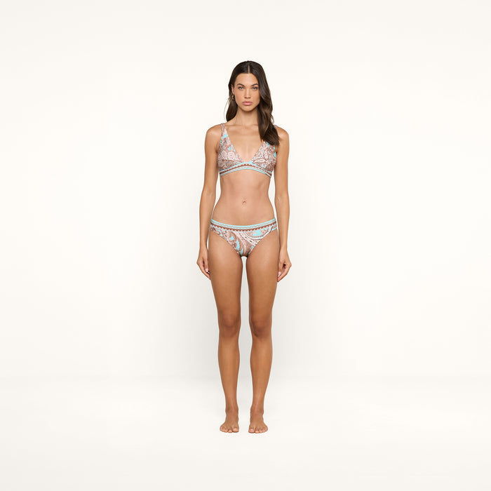 A woman stands barefoot against a plain white background, wearing the Lexie Bikini Top in a matching pastel paisley print. The bikini top features adjustable straps as she faces forward with a neutral expression and relaxed posture.