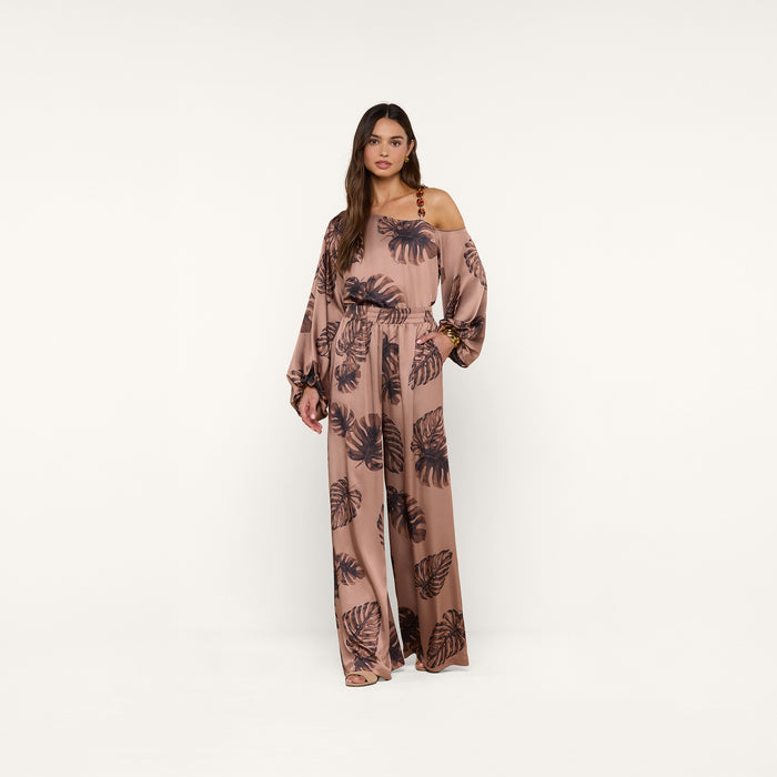 A woman stands against a plain white background, looking at the camera with one hand on her hip, wearing the Lillian Satin Pant in a loose fit with ultra-wide legs and a stylish black palm leaf print.
