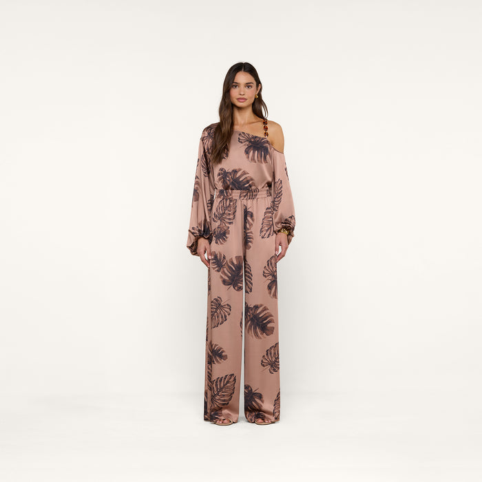 A woman models the Lillian Satin Pant, featuring an ultra-wide silhouette and a dark palm leaf print on pink satin. She stands against a plain white background, looking forward with a neutral expression.