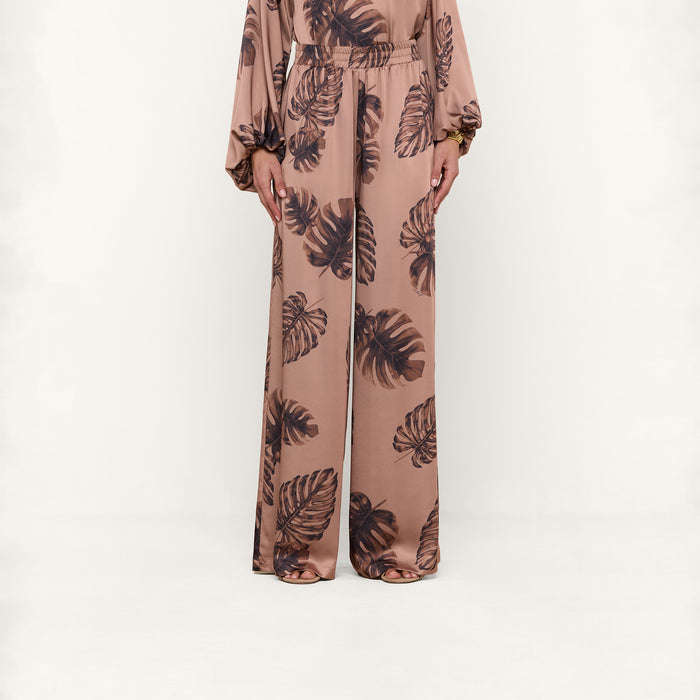 A person wearing the Lillian Satin Pant, featuring an ultra-wide leg and light brown satin fabric with a dark brown palm leaf print, stands against a plain white background; only their lower body is visible.