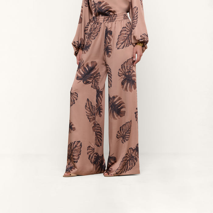 A person stands against a plain background, showcasing only the lower half of their outfit and wearing the Lillian Satin Pant featuring an ultra-wide silhouette with a brown palm leaf print.